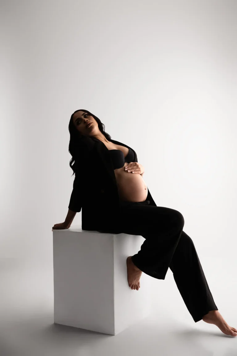 Pregnant woman seated on a white block in a dark studio, editorial portrait