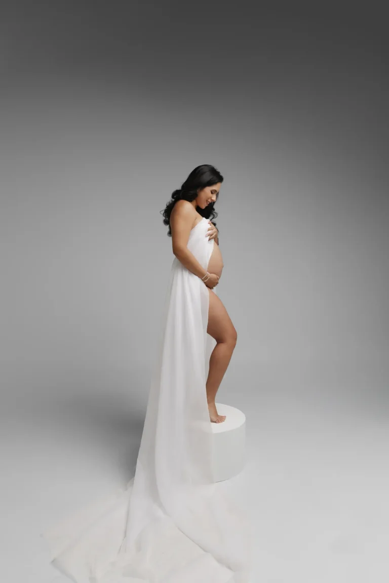 Pregnant woman in a flowing white gown standing in a studio, side profile