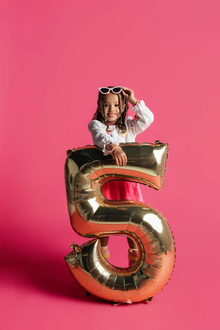 Child celebrating a fifth birthday with a gold number-five balloon on a pink studio backdrop
