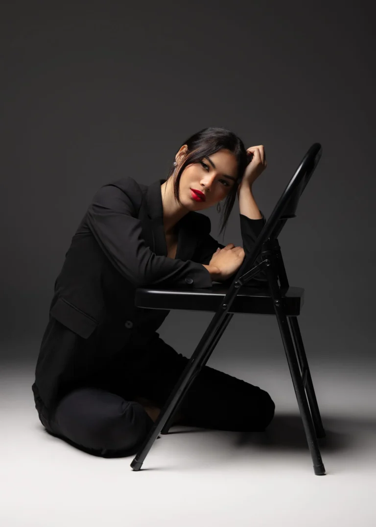 Woman in a black outfit seated on a chair in moody studio lighting